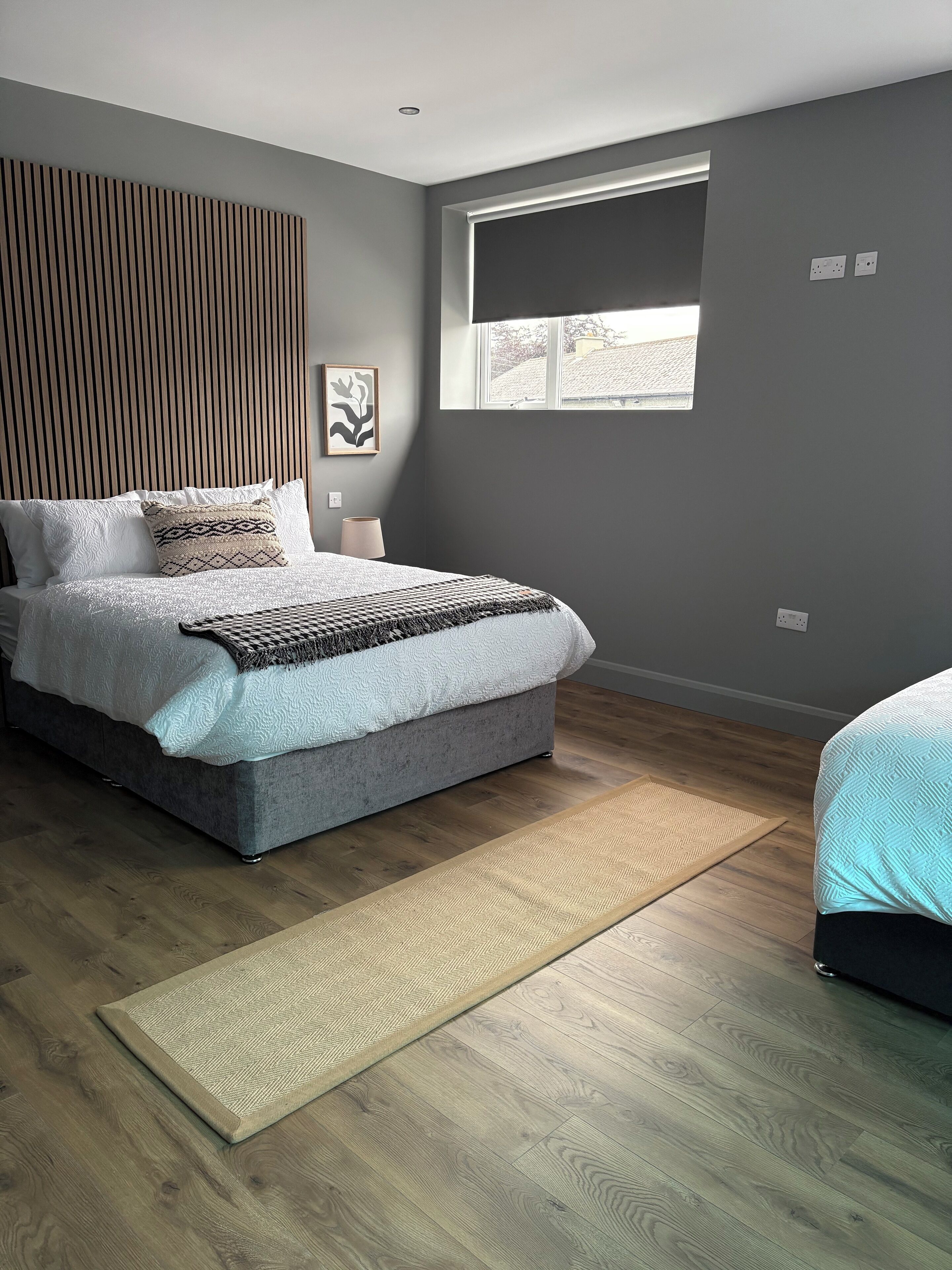 A modern bedroom with a grey bed, a wooden headboard, a large window, and a wooden floor.