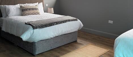 A modern bedroom with a grey bed, a wooden headboard, a large window, and a wooden floor.