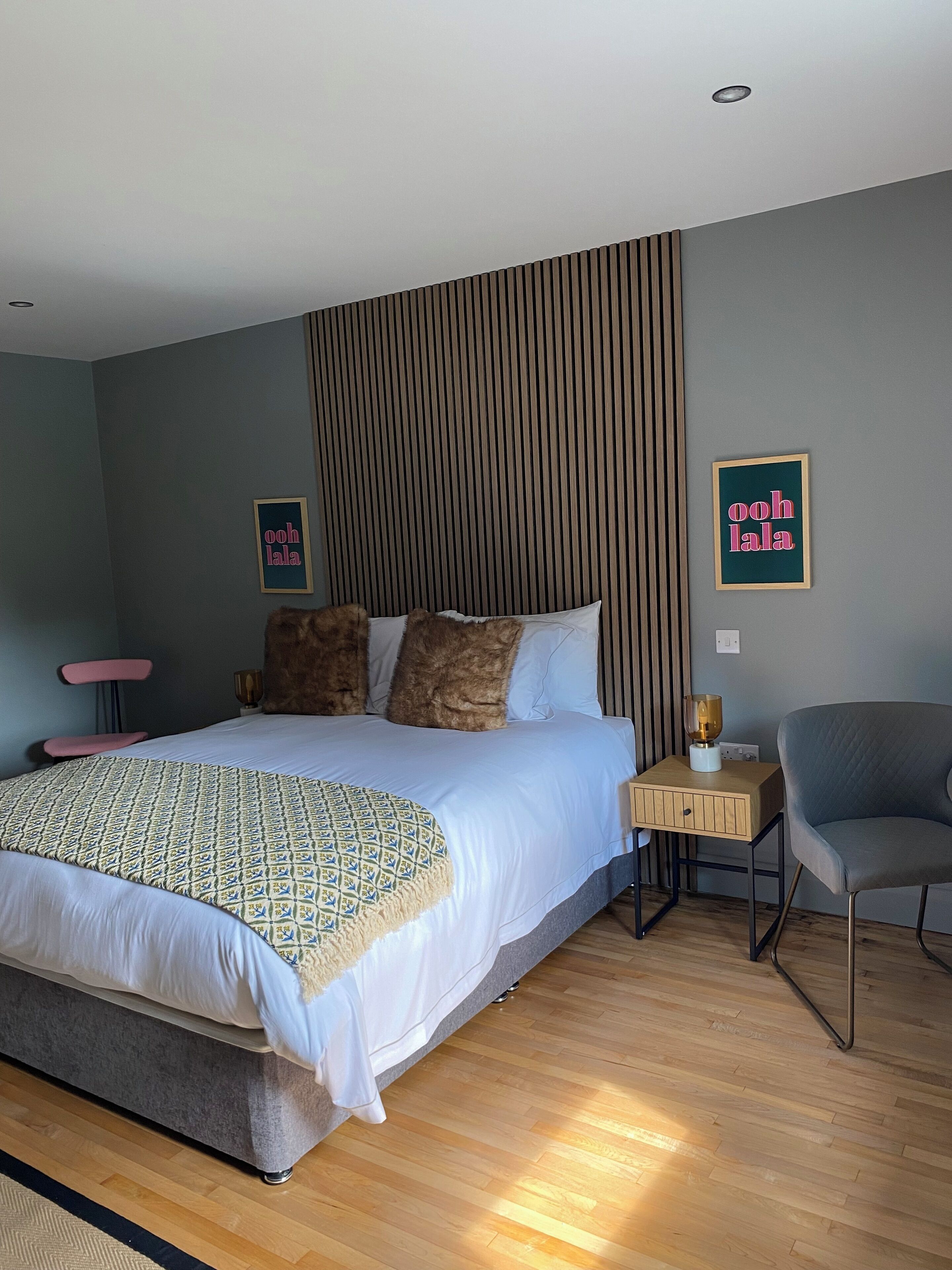 Un dormitorio moderno con una cama grande, una mesita de noche con lámpara, una silla y dos cuadros en la pared.
