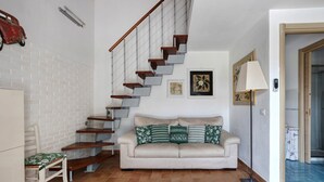 Ein Wohnzimmer mit Treppe, einer Couch mit gemusterten Kissen, einem Sessel, einer Lampe und einer Tür zu einem anderen Raum.