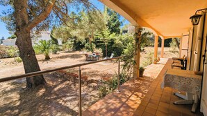 Une terrasse extérieure couverte, dotée d’un sol en pierre, d’une rambarde en métal et offrant une vue sur un jardin avec des arbres et une table entourée de chaises.