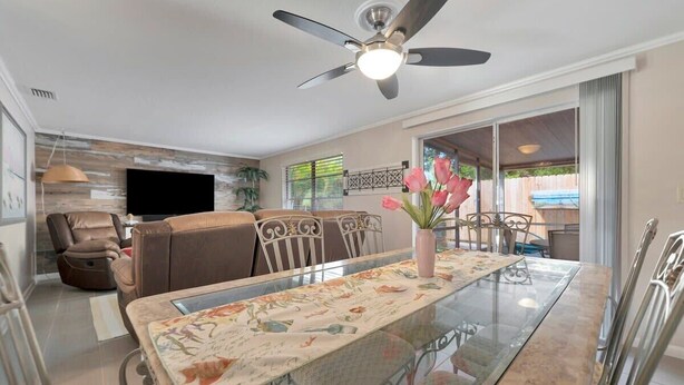 A spacious living room with a glass-top dining table, a ceiling fan, a sofa, and a TV mounted on the wall.