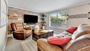 A living room with a leather sofa, a recliner, a flat-screen TV, a coffee table, and a large window with blinds.