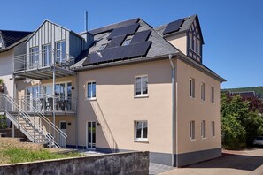 Ein mehrgeschossiges Gebäude mit einem Balkon, Solaranlage auf dem Dach und einer Treppe, die zu den oberen Stockwerken führt.
