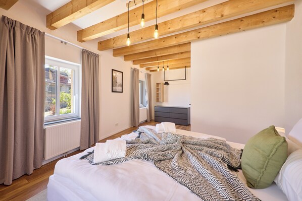 Ein Schlafzimmer mit Holzboden, einem Bett mit gemusterten Betttextilien, einem Schminktisch und einem Fenster mit Vorhängen.