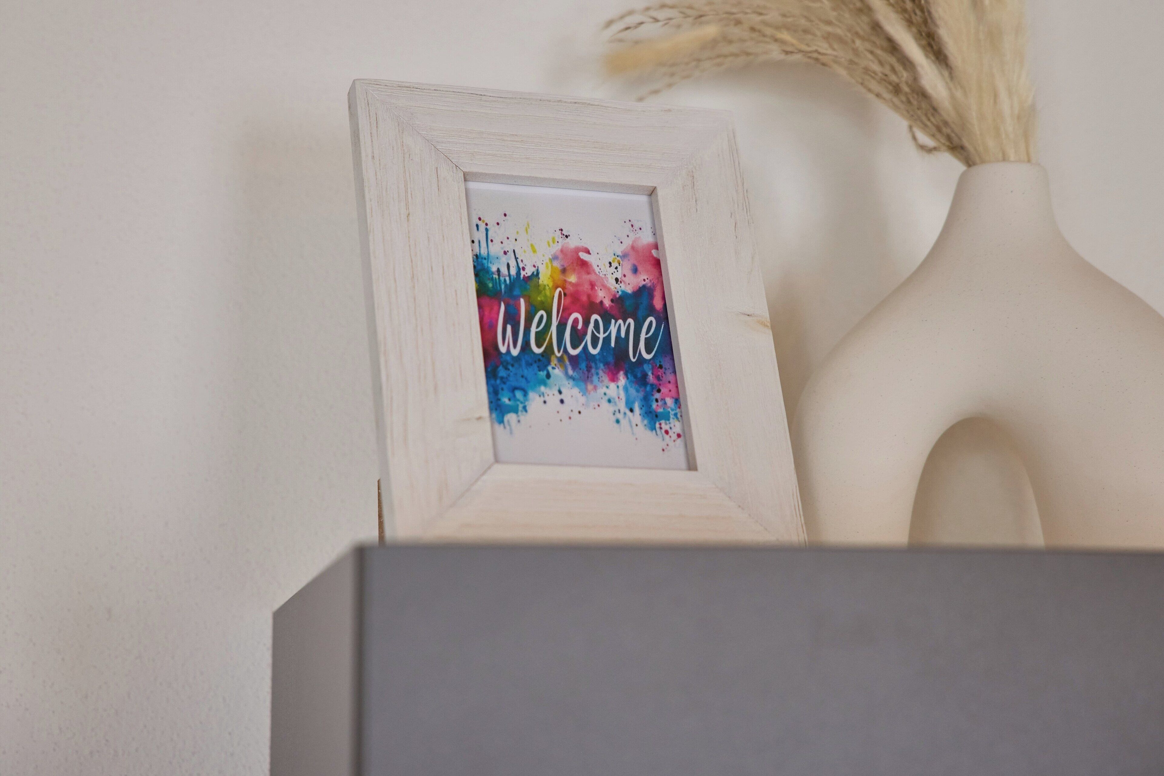 A framed welcome sign with colorful splashes of paint and the word 'Welcome' in cursive.
