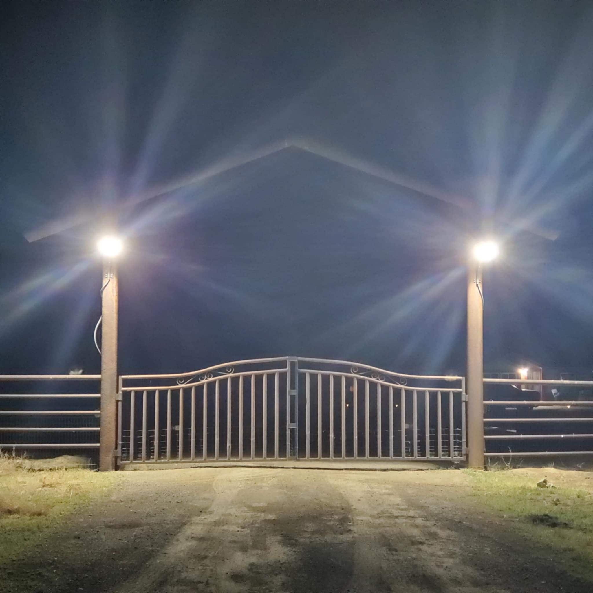 Une entrée fermée par un portail, avec deux lampadaires, un portail en métal et un chemin de terre menant à un bâtiment.