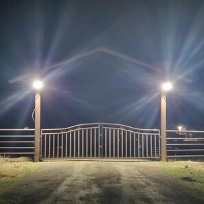 Portão fechado com dois postes de luz, um portão de metal e um caminho de terra que leva a um edifício.
