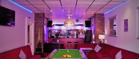 A billiards room with a pool table, red sofas, a bar area, and a chandelier.
