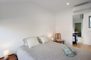 Ein Schlafzimmer mit einem Bett, Kissen, einem Nachttisch mit Lampe, einem Sessel und einer Klimaanlage.
