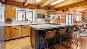 A log cabin kitchen with wooden cabinets, a large island with bar stools, a microwave, and a large window. - Lodge Hot Tub Dueling Basketball (Park City)