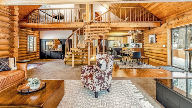 A spacious log cabin interior with a central staircase, a cozy seating area, and a kitchen visible in the background. - Lodge Hot Tub Dueling Basketball (Park City)