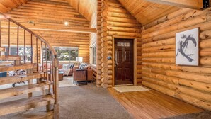 A log cabin interior with a wooden staircase, a living area with a sofa and coffee table, and a large wooden door. - Lodge Hot Tub Dueling Basketball (Park City)