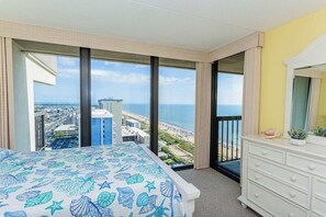 Ein Hotelzimmer mit einem großen Bett, einem Schminktisch und Blick auf Strand und Stadt.