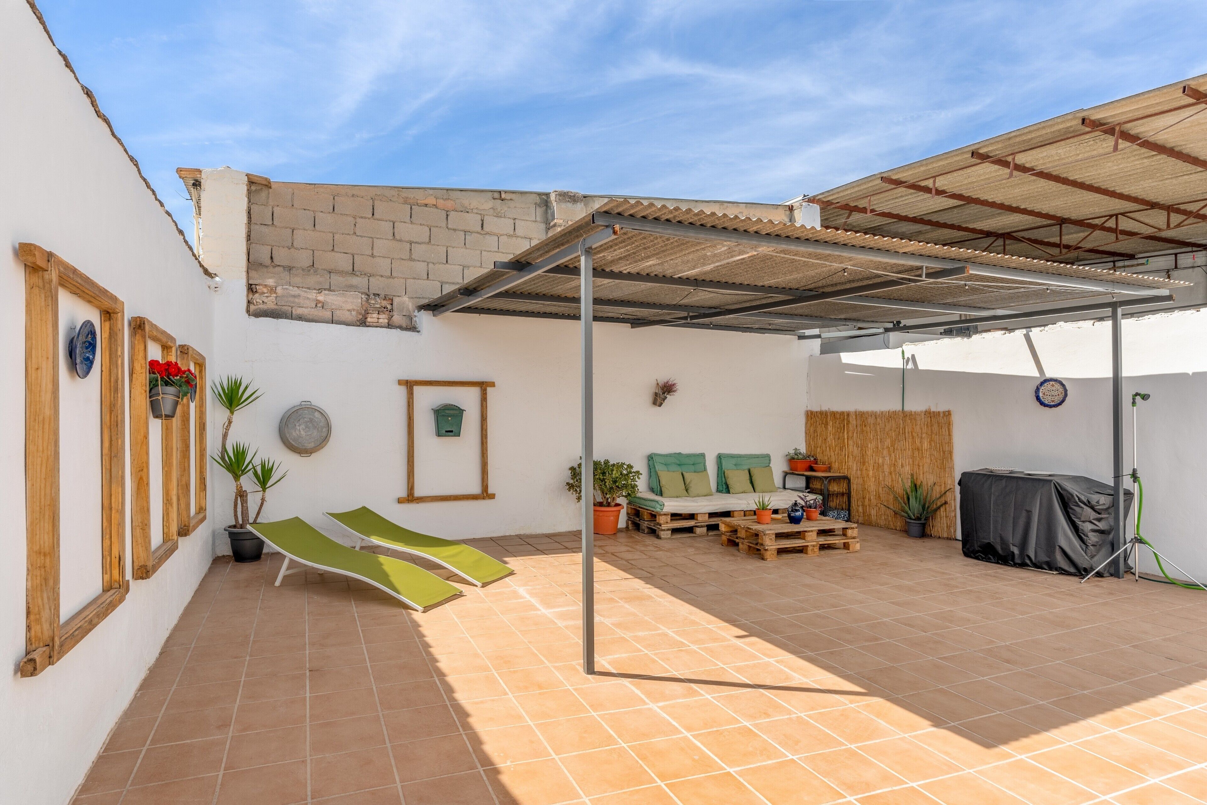 Ein überdachter Patio-Bereich mit Fliesenboden, einem grünen Lounge-Sessel, einer Holzbank und Topfpflanzen.