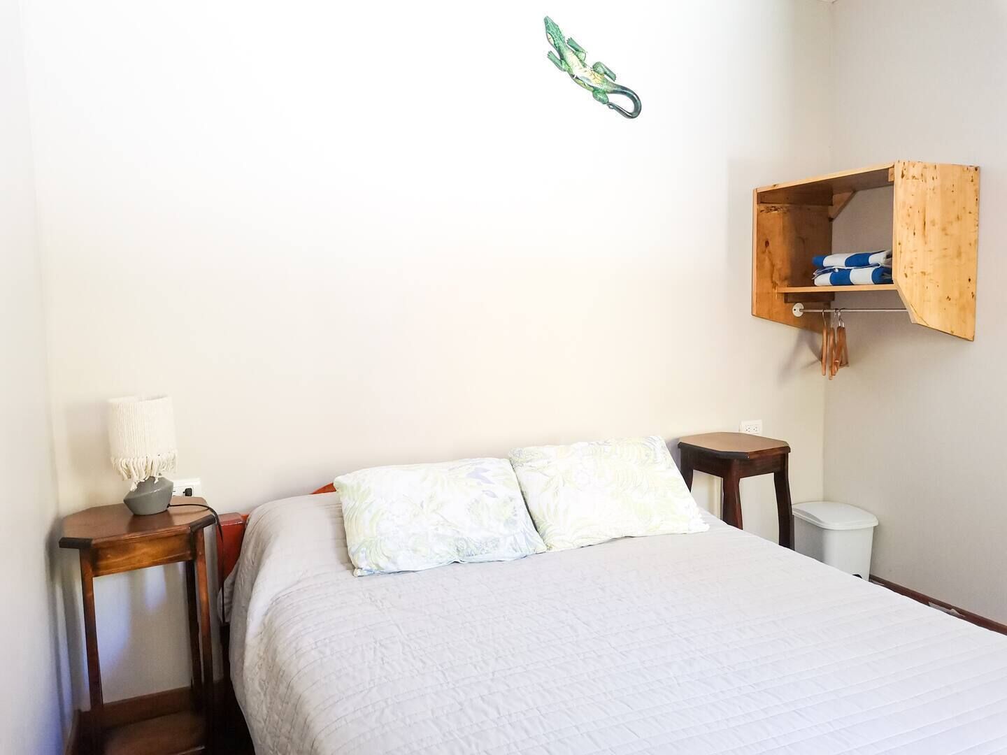 Een slaapkamer met een bed, een nachtkastje, een lamp, een houten plank en een haak aan de muur.