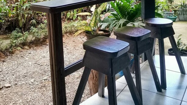 Ein rustikaler Sitzbereich im Freien mit einem Holztisch und Hockern, umgeben von üppigem Grün und Pflanzen.