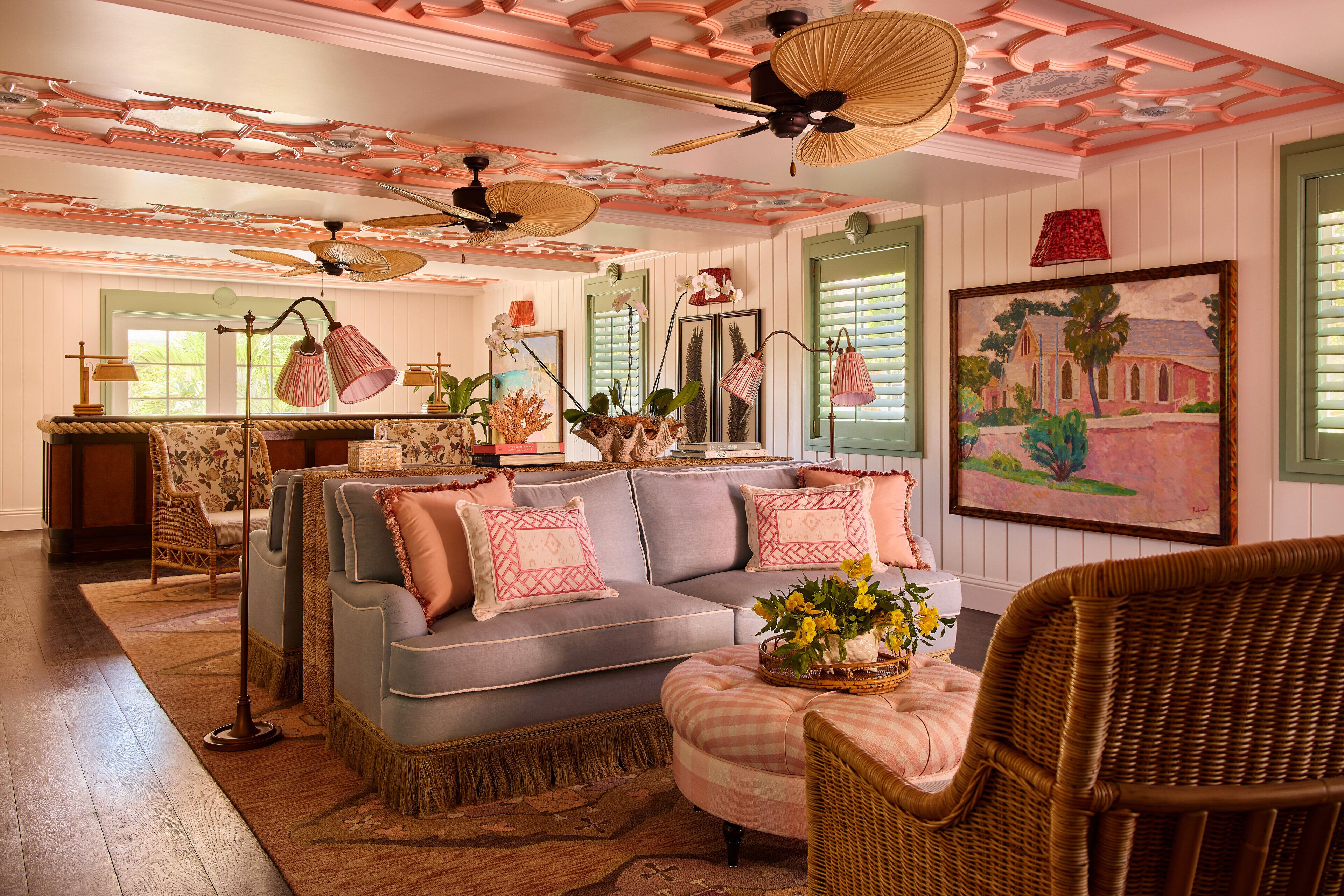 A room with a grey sofa, wicker chairs, a ceiling fan, and a painting on the wall.