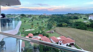Un balcón con vistas a un campo de golf, una zona residencial con casas y una costa distante.