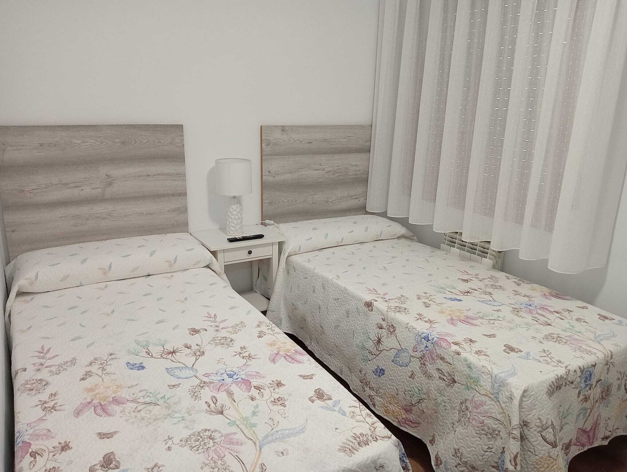 Une chambre avec deux lits, chacun agrémenté de draps à motifs floraux, une table de chevet avec une lampe et une fenêtre ornée de rideaux légers.