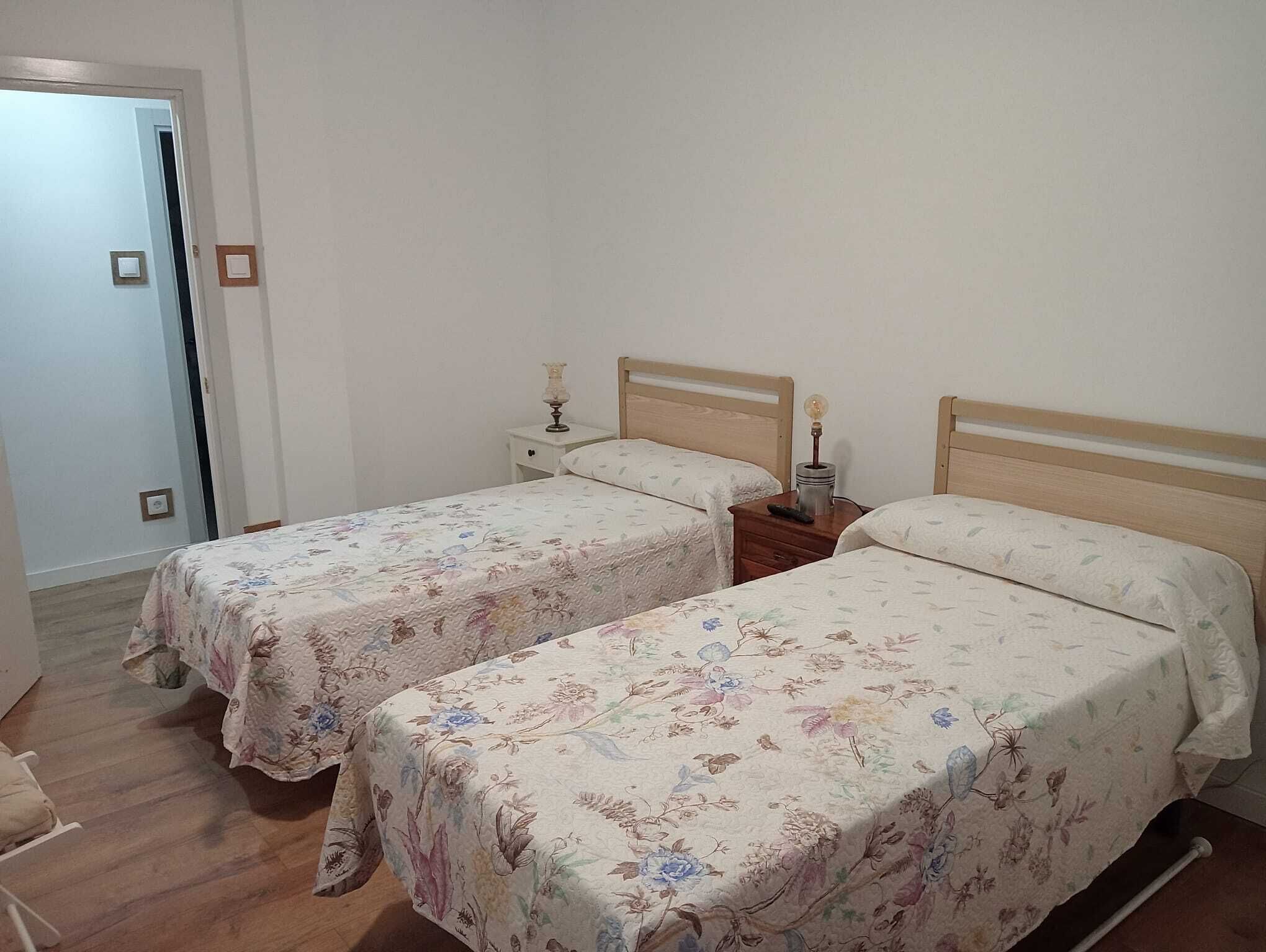 Quarto com duas camas de solteiro, cada uma com cabeceira de madeira e mesa de cabeceira com abajur.