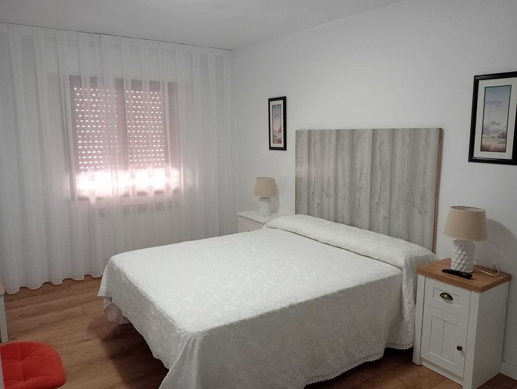 A bedroom with a bed, a nightstand, a lamp, a red chair, and a window with curtains.