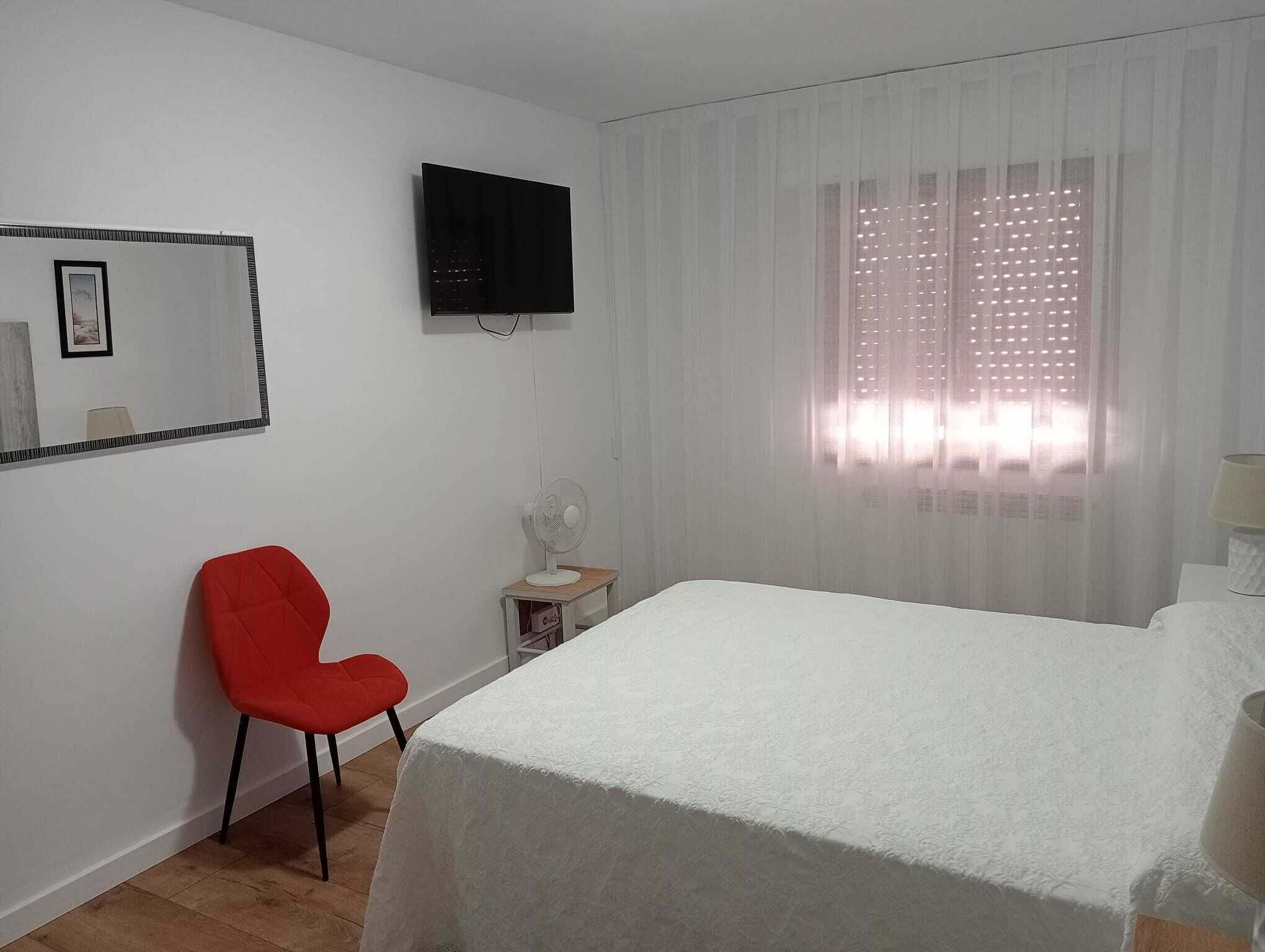 Ein Hotelzimmer mit Bett, einem roten Sessel, einem kleinen Tisch mit Lampe, einem an der Wand montierten Fernseher und einem Fenster mit durchsichtigen Vorhängen.