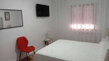 Ein Hotelzimmer mit Bett, einem roten Sessel, einem kleinen Tisch mit Lampe, einem an der Wand montierten Fernseher und einem Fenster mit durchsichtigen Vorhängen.