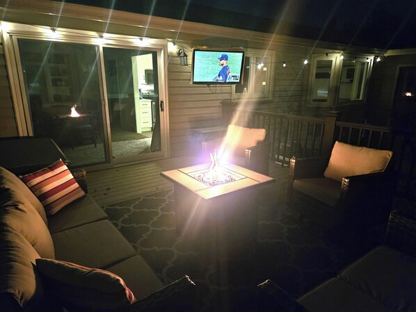 Un patio al aire libre con una televisión instalada en una pared, un elemento decorativo de fuego y arreglos de asientos.