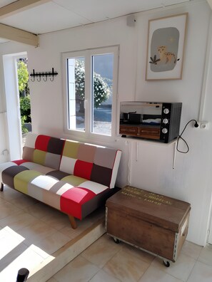 Una habitación con un sofá colorido, un cofre de madera, un radio instalado en la pared y una ventana con vistas a follaje.
