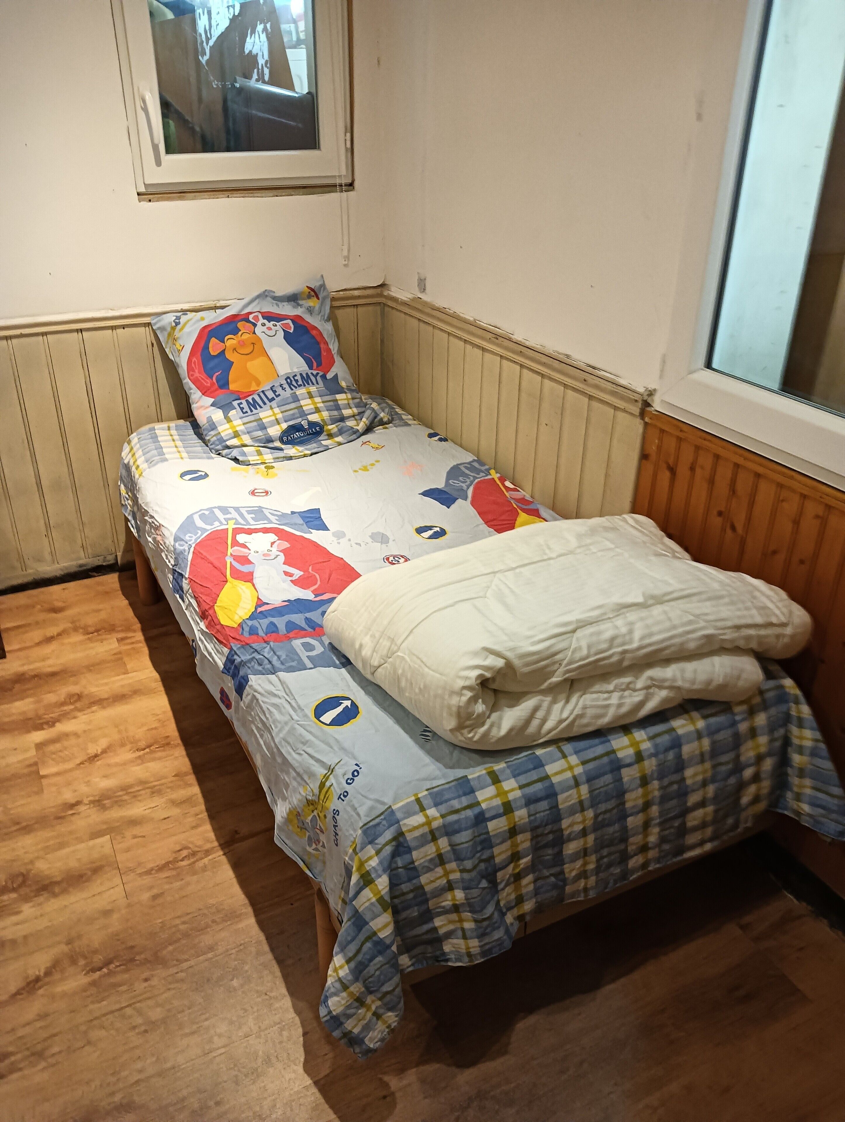 Un lit simple avec une couvre-lit aux couleurs vives, un oreiller orné d’un personnage de dessin animé et une fenêtre au-dessus du lit.