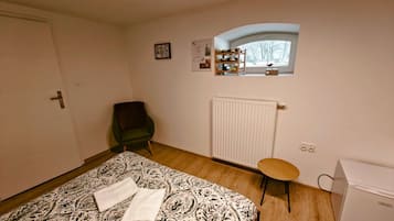 Un dormitorio con una cama, una silla, una mesita, un radiador, una ventana y un refrigerador.