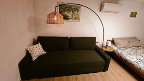 Een gezellige kamer met een donkergroene bank, een kussen met patroon, een rieten lamp, een bed met beddengoed met dessin, en een wandairconditioner.