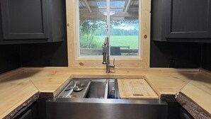 A kitchen with dark cabinets, a wooden countertop, a stainless steel sink, and a window overlooking a green landscape.