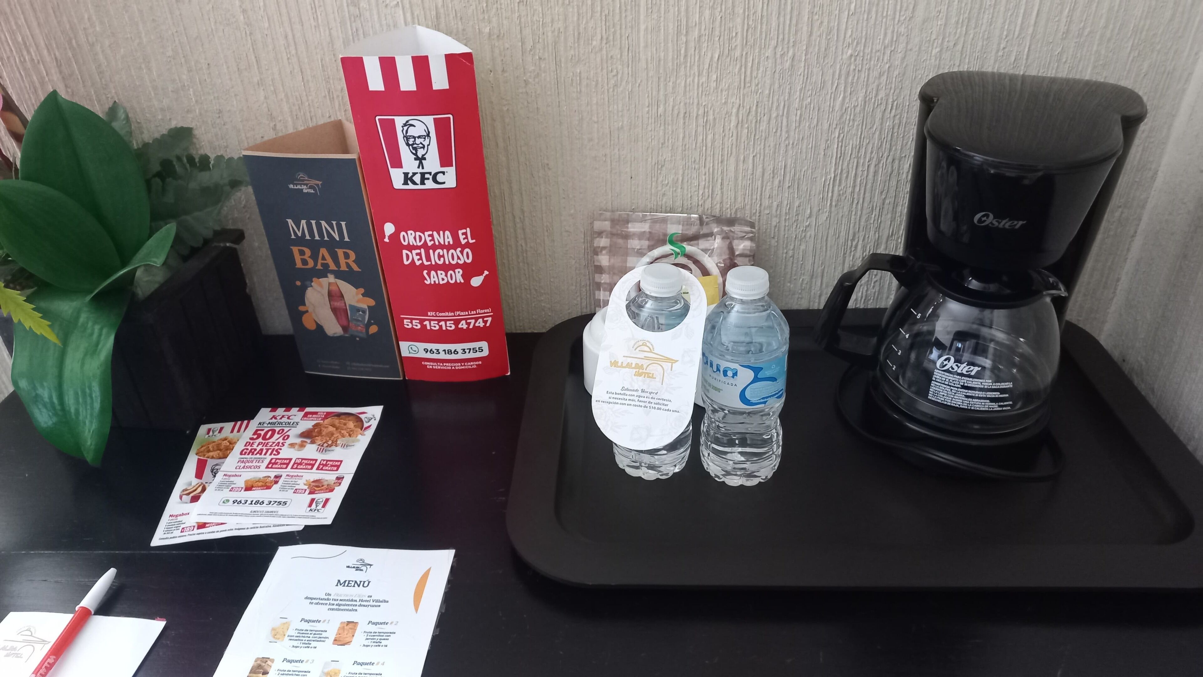 A hotel room with a KFC mini bar, a menu, a water bottle, and a coffee maker on the table.