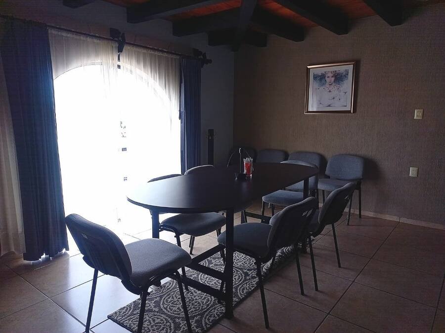 A dining area with a round table and six chairs, a framed picture on the wall, and a window with curtains.