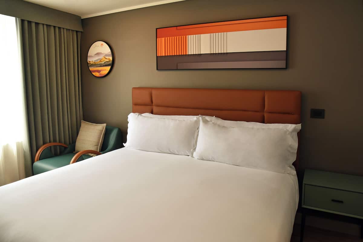 A hotel room with a large bed, a headboard, a nightstand, a chair, and a wall art piece.