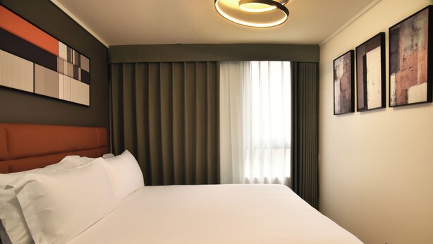 A hotel room with a large bed, a headboard, a window with curtains, and wall-mounted artwork. - APART HOTEL HW COMPAÑIA (Santiago)