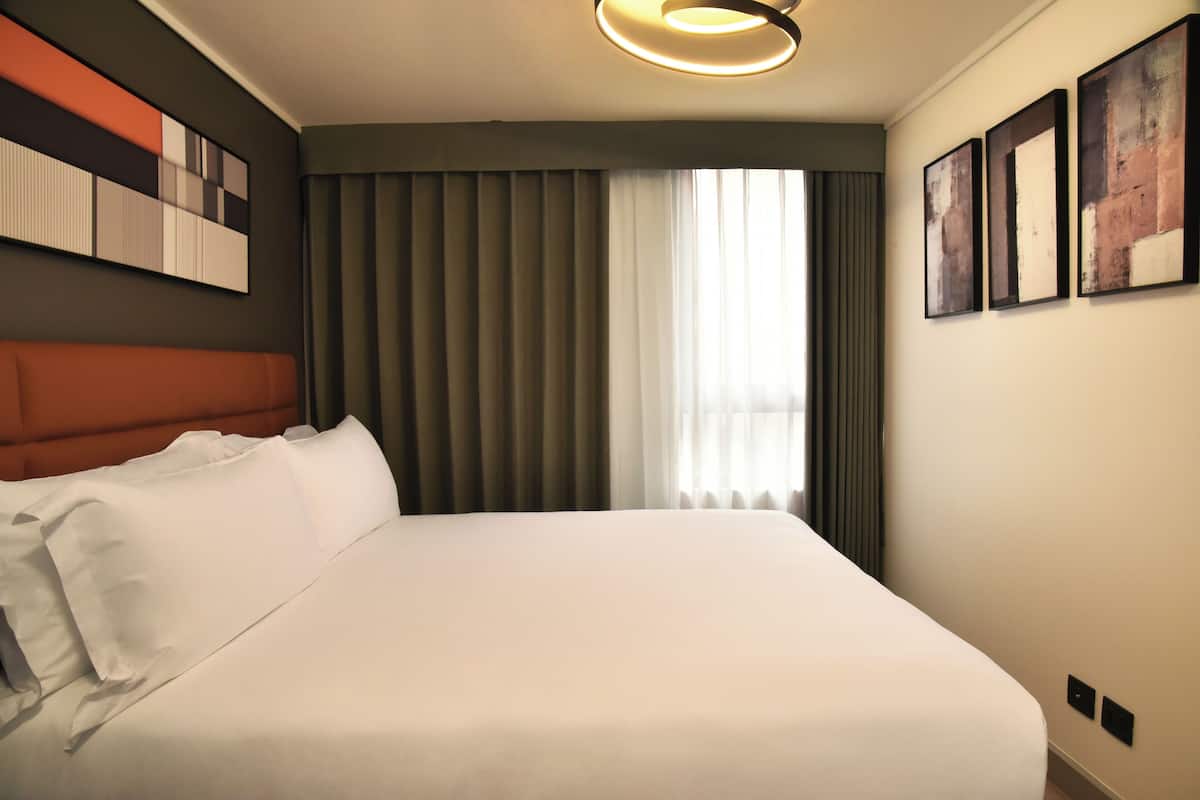 A hotel room with a large bed, a headboard, a window with curtains, and wall-mounted artwork.