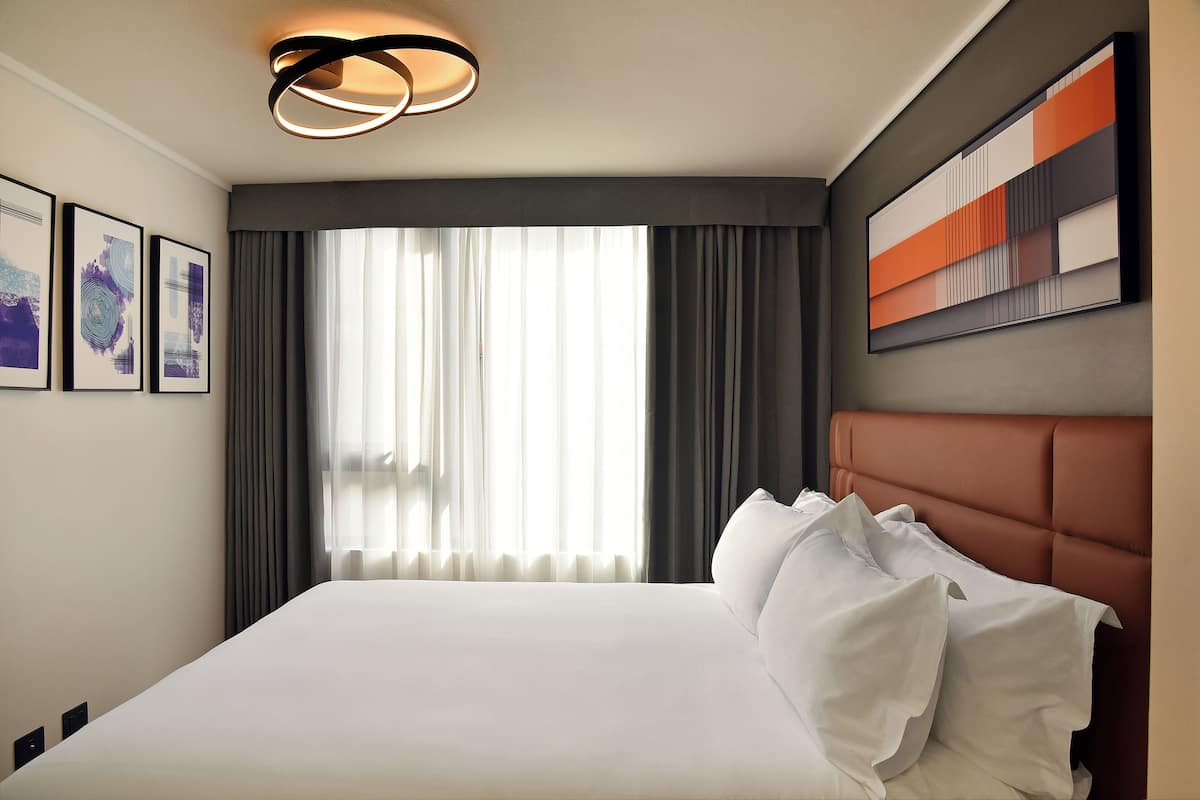 A hotel room with a large bed, a modern light fixture, a large window with curtains, and a piece of abstract art on the wall.