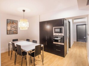 A modern kitchen with dark cabinetry, a built-in microwave, and an oven. There is a dining table with chairs and a pendant light. A framed picture hangs on the wall.