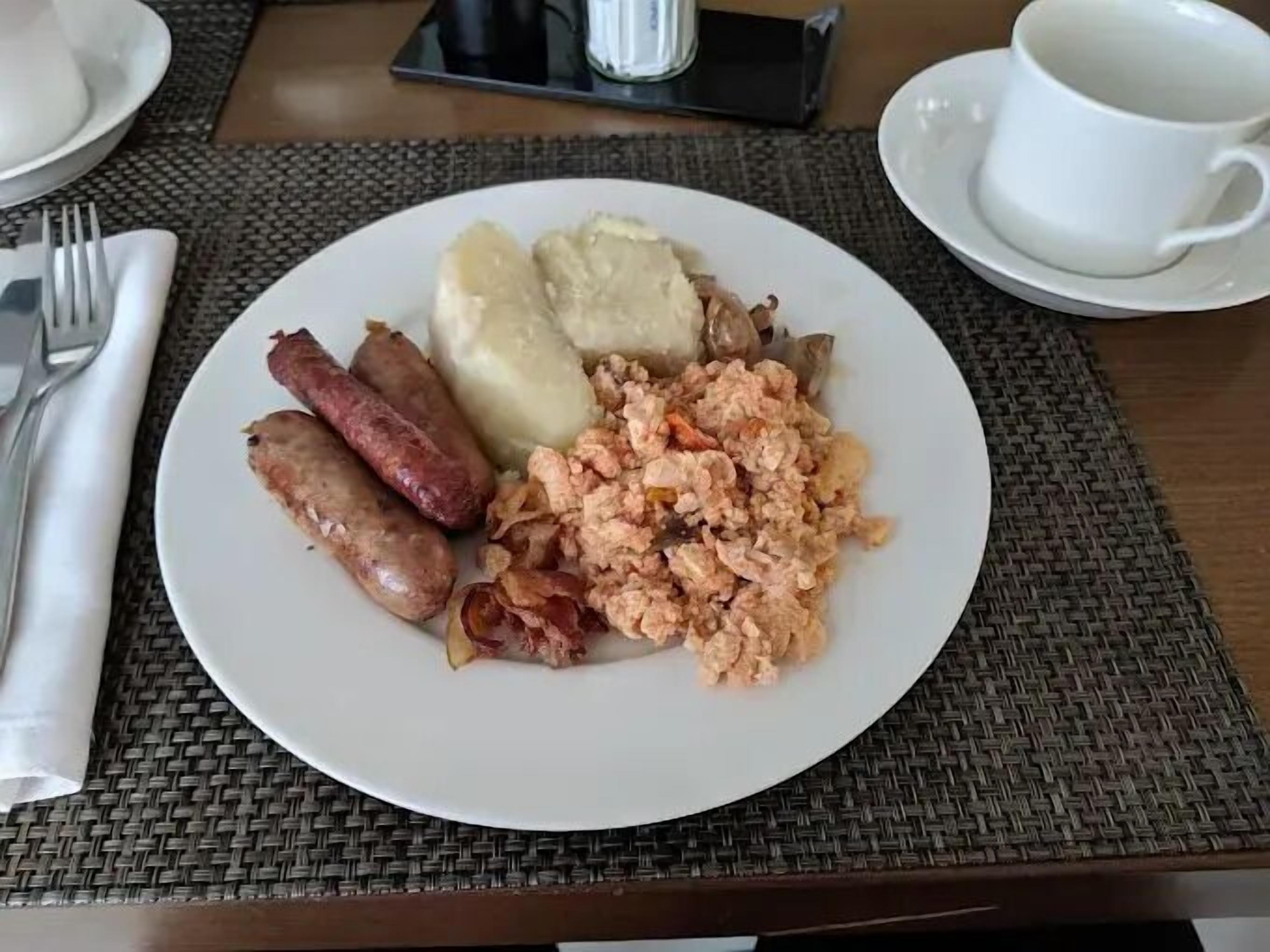 A breakfast plate with sausages, mashed potatoes, and scrambled eggs on a woven placemat.