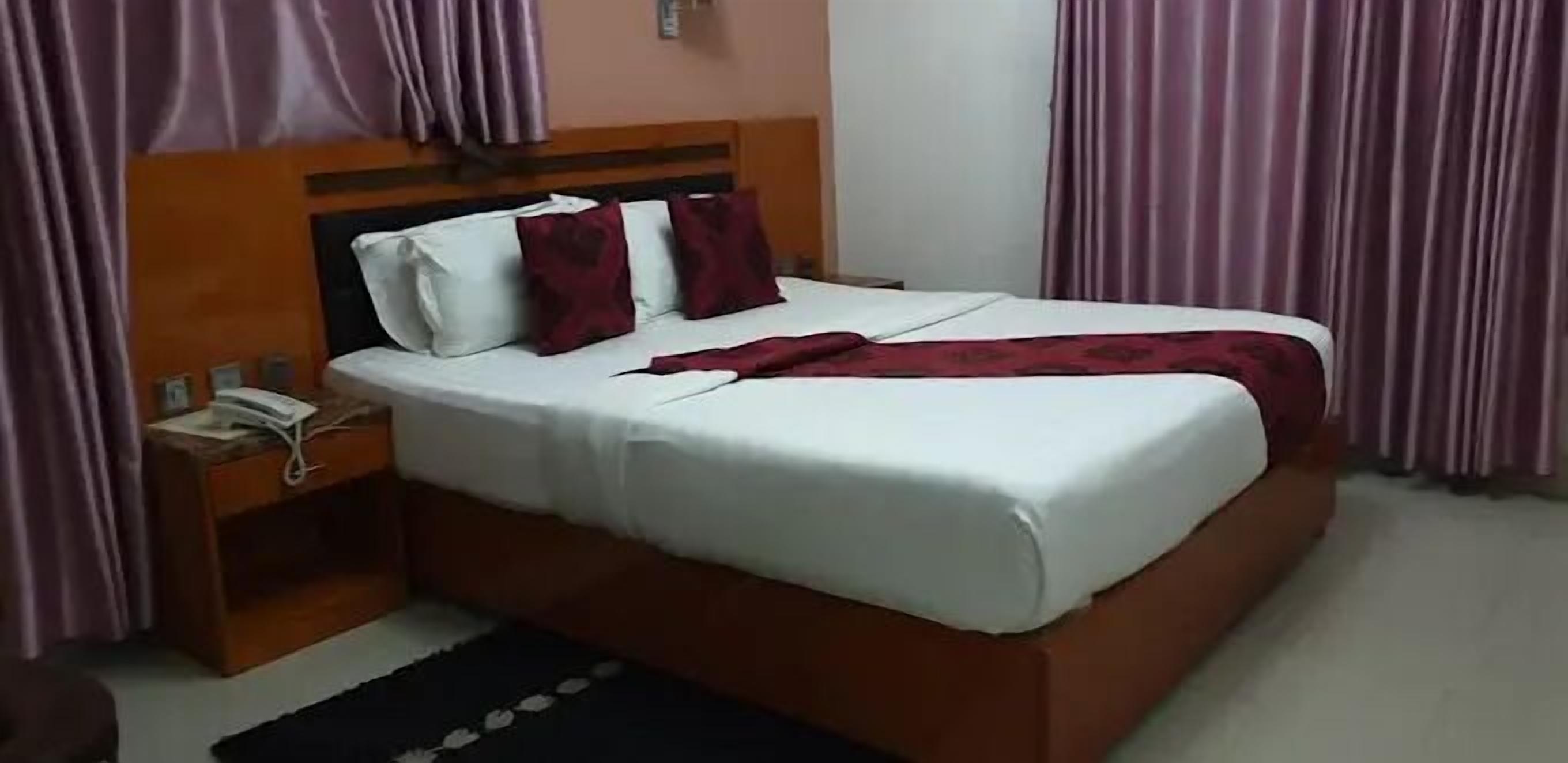A neatly made bed with white linens and a red blanket, a wooden nightstand with a telephone, and purple curtains.