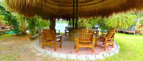 A thatched gazebo with wooden chairs and a table, surrounded by lush greenery.