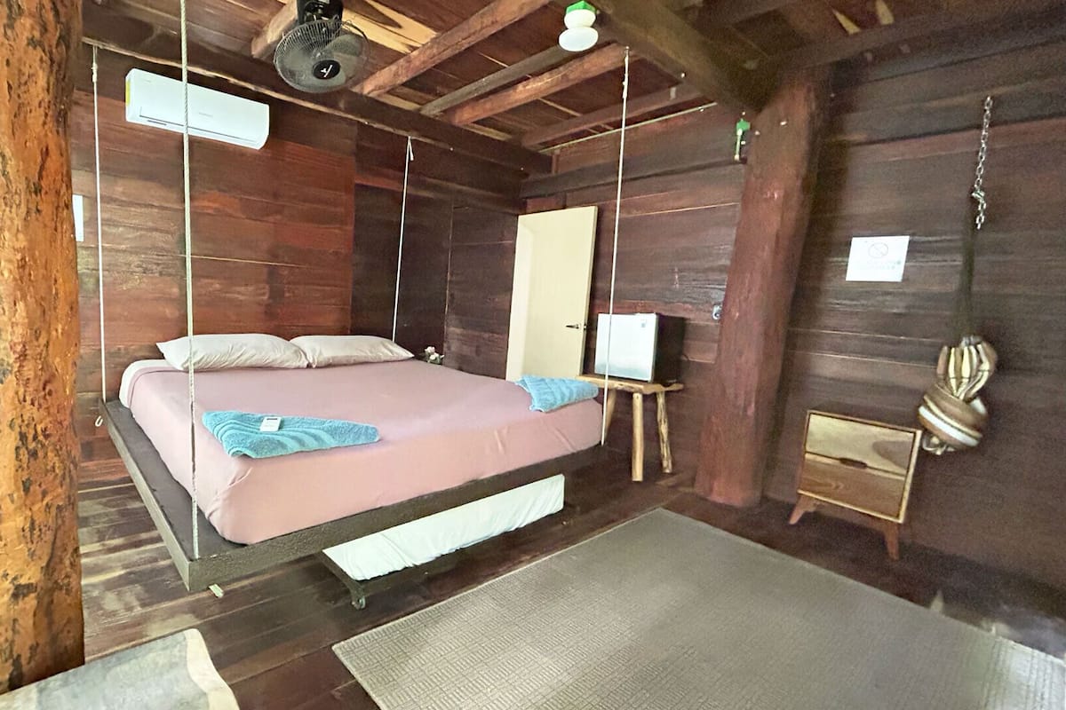 A rustic wooden room with a suspended bed, a small bedside table, a mirror, and a decorative item on the wall.