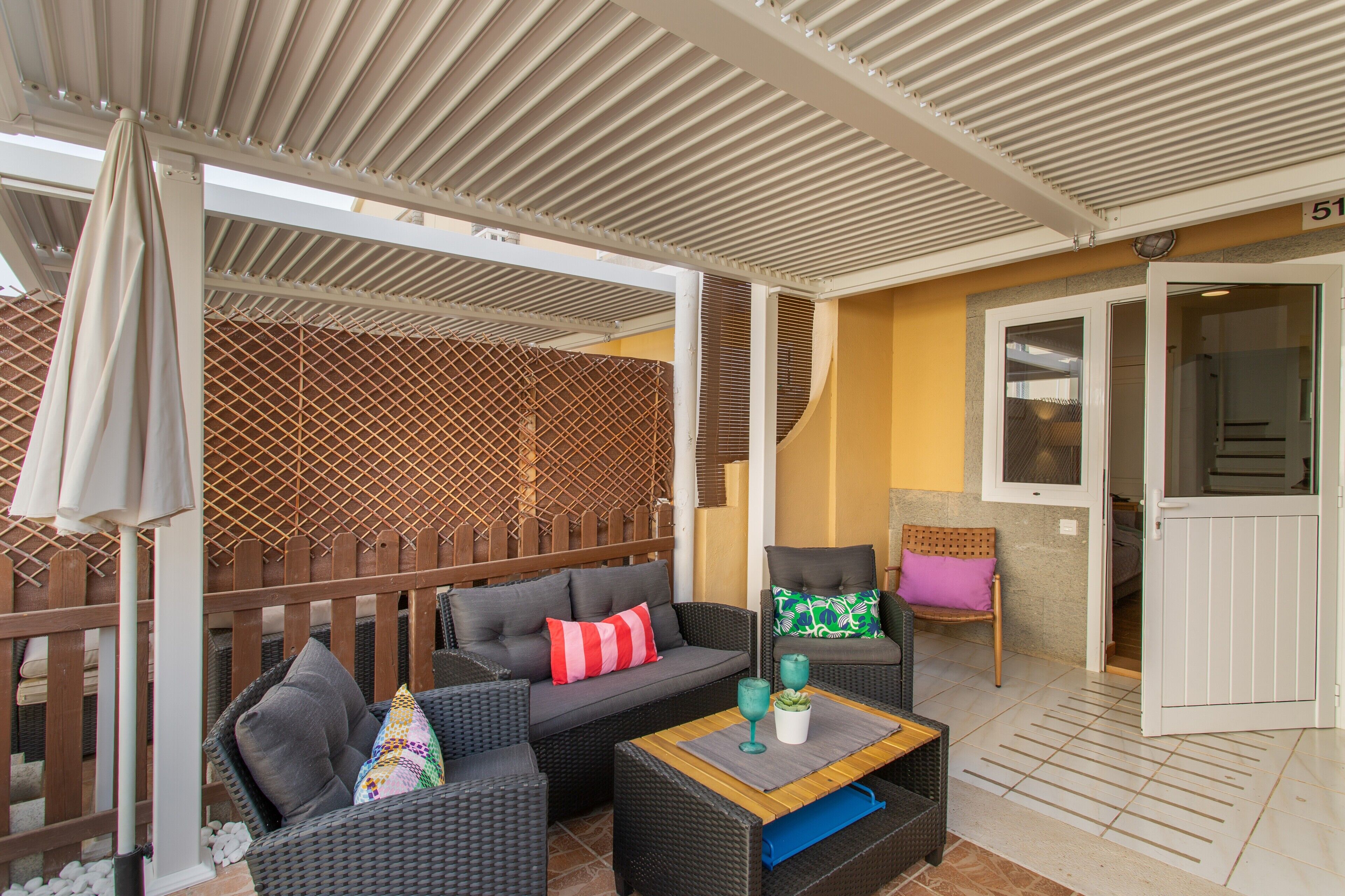 A covered outdoor seating area with wicker furniture, a wooden fence, and a door leading to an interior room.