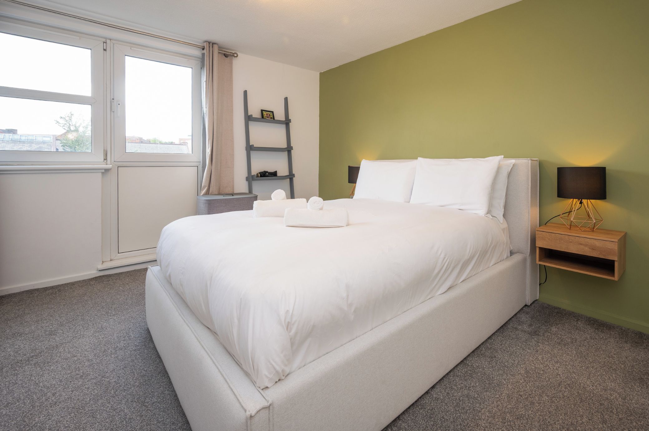 A hotel room with a bed, a nightstand with a lamp, a window with curtains, and a shelf with a picture frame.