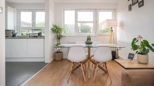 A modern kitchen with white cabinets, a glass-top dining table, two chairs, a potted plant, and a view of the outside through large windows. - Sun Filled 1BD Split Level Flat by Caledonian Road (Greater London)