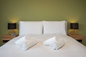 A neatly made bed with white linens and two rolled towels, flanked by two bedside tables with lamps, against a green wall.