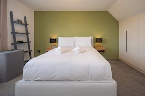 Ein ordentlich bezogenes Bett mit weißen Leinen, zwei Nachttischlampen, ein kleines Regal und ein grauer Nachttisch in einem Zimmer mit grünen Wänden.